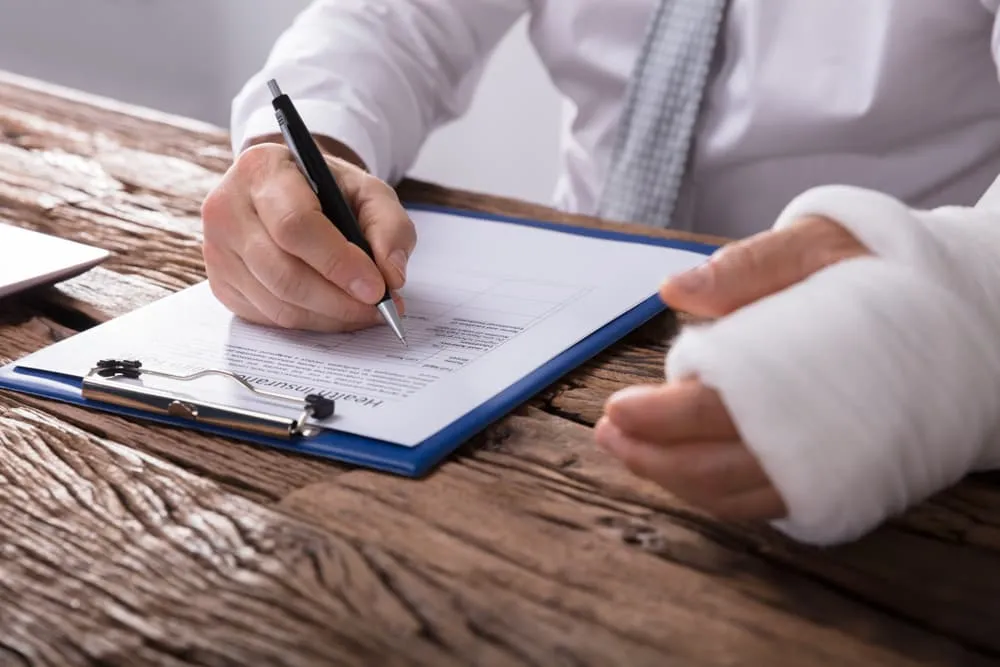 A person with a bandaged hand fills out a health insurance form on a clipboard at a wooden table.