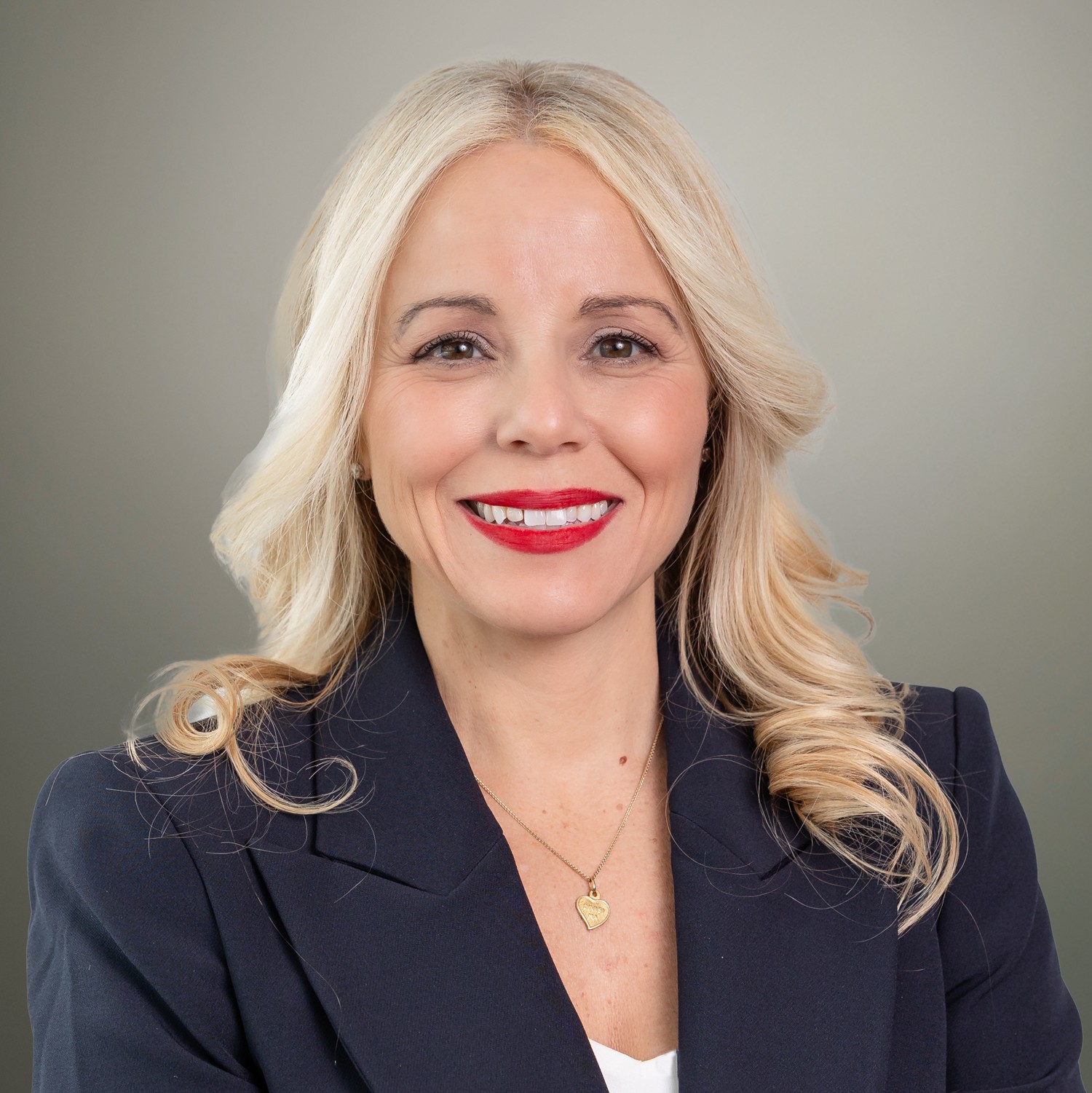 A woman with long blonde hair, wearing a navy blazer and red lipstick, smiles at the camera against a plain background.