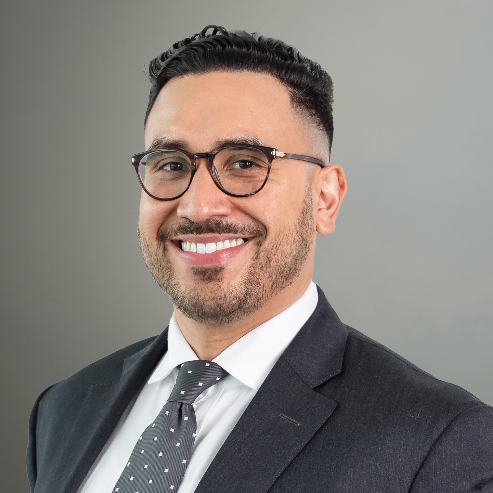 Man with short dark hair, beard, and glasses wearing a dark suit, white shirt, and patterned tie, smiling in front of a plain gray background.