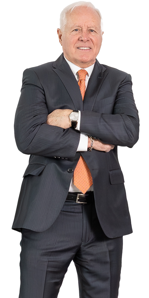 Older man in a dark suit and orange tie stands with arms crossed, looking forward with a neutral expression against a plain background.