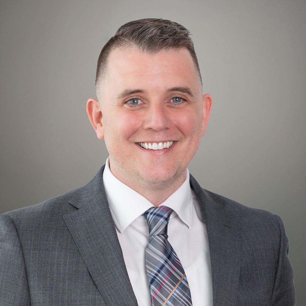 A man in a gray suit and patterned tie smiles at the camera against a plain gray background.