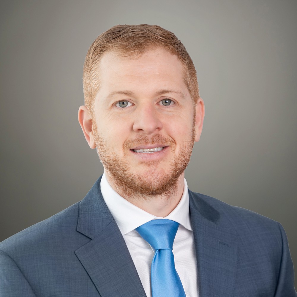 A man with short light brown hair and a beard, wearing a blue suit, white shirt, and blue tie, poses against a neutral gray background.