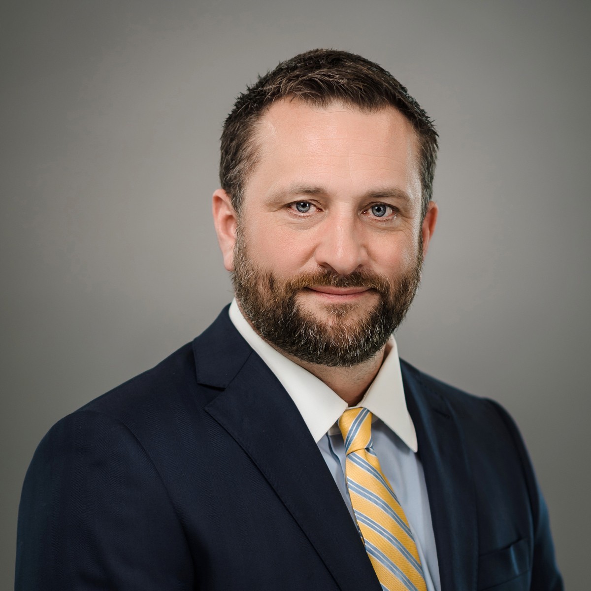 A man with short brown hair and a beard wearing a navy suit, white shirt, and yellow striped tie poses against a plain grey background.