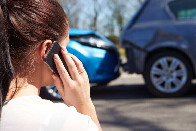 A woman is making a phone call while standing near two cars involved in a rear-end collision on a road.