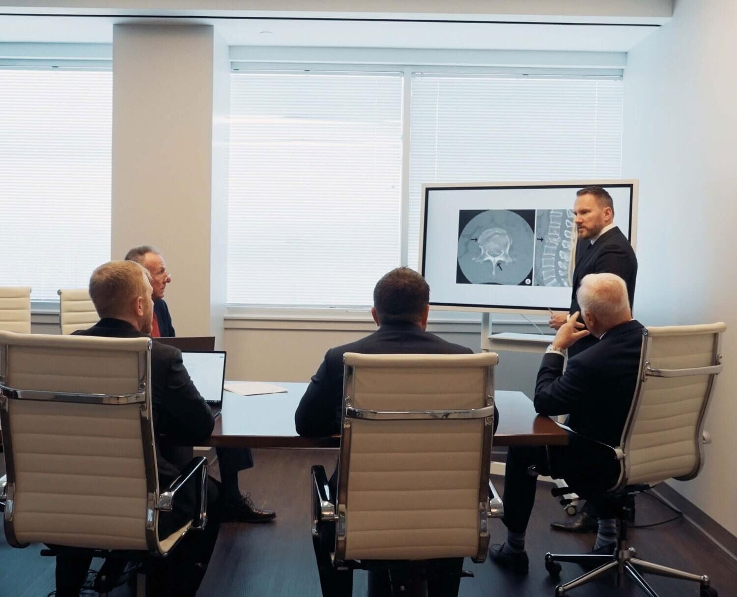 Four men in business suits sit around a conference table as another man presents a medical scan displayed on a large screen at the front of the room.