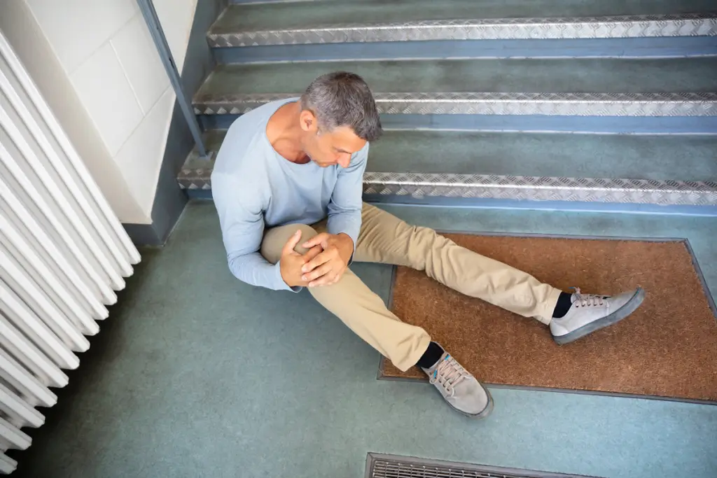 A man sits on the floor at the base of stairs, holding his knee and appearing to be in pain, suggesting he may have fallen.